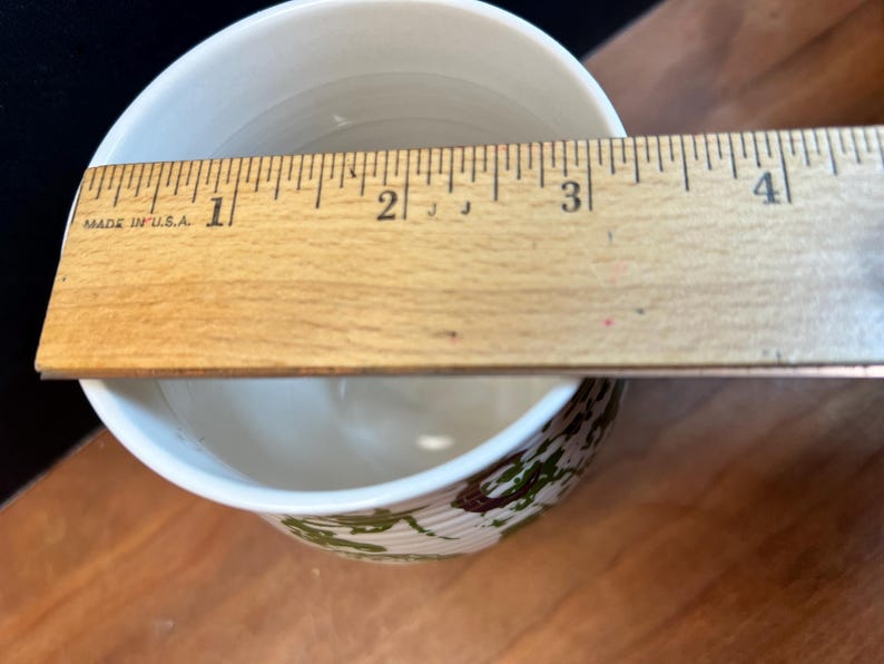 May include: A white ceramic bowl with a decorative green and brown pattern around the base. A wooden ruler, marked in inches, is placed across the top of the bowl, measuring approximately 3 inches in diameter.