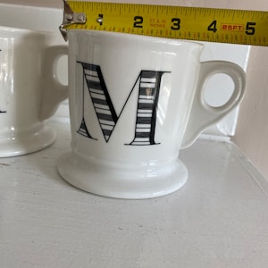 Puede incluir: Taza de cer&aacute;mica blanca con un dise&ntilde;o de la letra "M" en negro. La taza tiene un asa y una base con pie. La taza mide aproximadamente 9 cm de alto, medidos con una cinta m&eacute;trica amarilla.