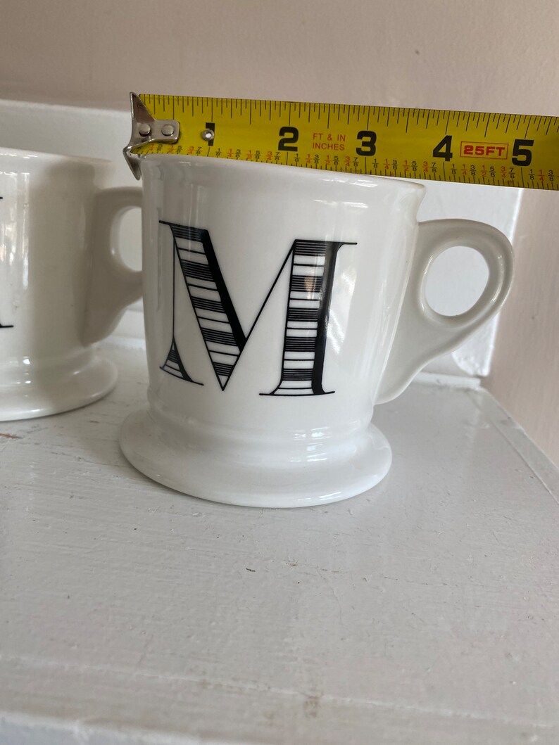 Puede incluir: Taza de cer&aacute;mica blanca con un dise&ntilde;o de la letra "M" en negro. La taza tiene un asa y una base con pie. La taza mide aproximadamente 9 cm de alto, medidos con una cinta m&eacute;trica amarilla.