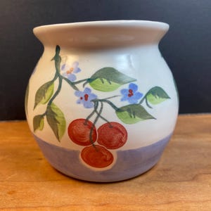 May include: A small, hand-painted ceramic vase with a white upper section and a lavender-blue base. The vase is decorated with a cherry branch design featuring red cherries, green leaves, and blue flowers. The vase is likely used for decorative purposes.