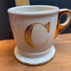 May include: White ceramic mug with a gold-coloured handle and a gold-coloured letter "C" on the front.