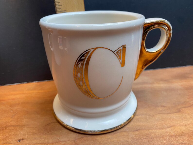 May include: White ceramic mug with a gold-coloured handle and a gold-coloured letter "C" on the front.