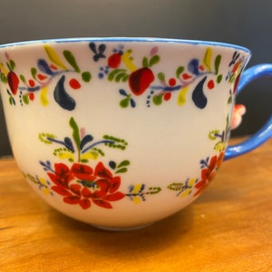 K&ouml;nnte beinhalten: Wei&szlig;e Keramik-Teetasse mit blauem Henkel und Rand, verziert mit einem Blumenmuster in Rot, Gelb und Blau. Die Tasse hat ein gro&szlig;es rotes Blumendesign und einen kleinen floralen Akzent am Henkel.