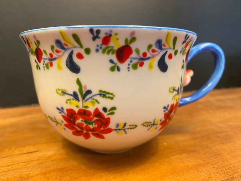 K&ouml;nnte beinhalten: Wei&szlig;e Keramik-Teetasse mit blauem Henkel und Rand, verziert mit einem Blumenmuster in Rot, Gelb und Blau. Die Tasse hat ein gro&szlig;es rotes Blumendesign und einen kleinen floralen Akzent am Henkel.