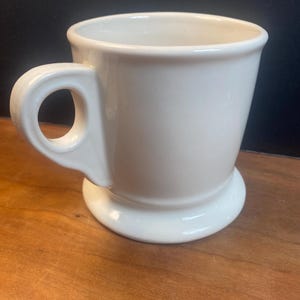 May include: A white ceramic mug with a rounded handle and a wide, sturdy base. The mug has a simple, classic design and a smooth, glossy finish. The mug is empty and ready to be filled with a beverage.