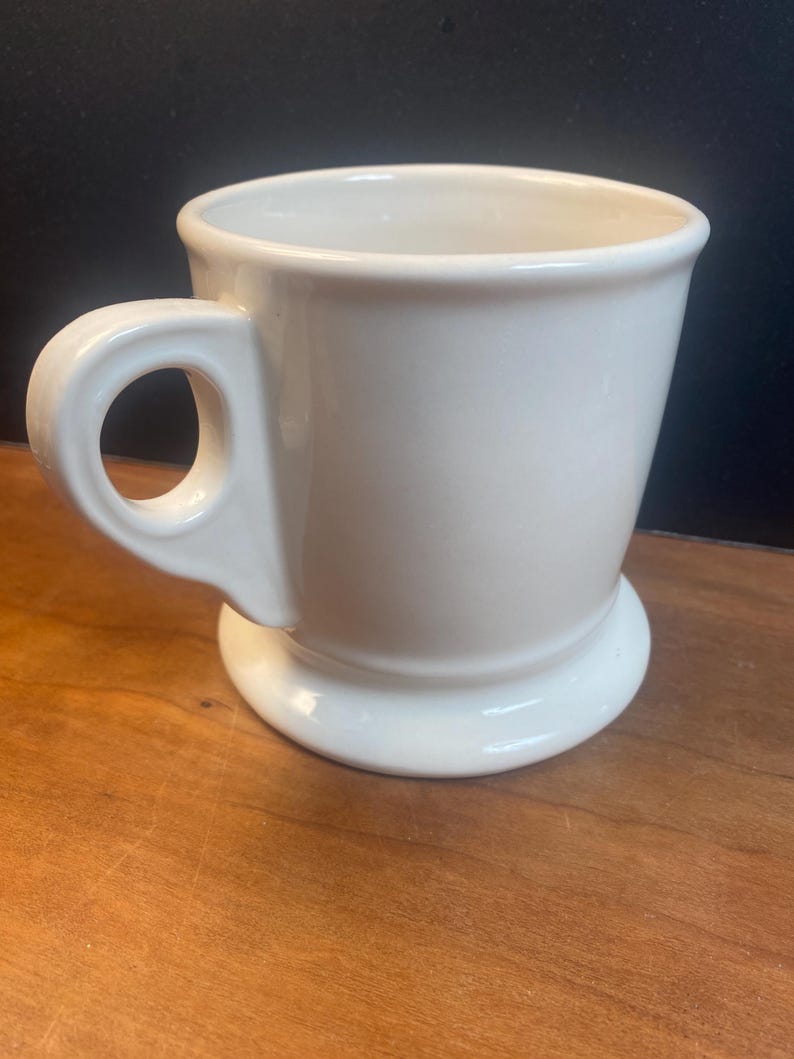 May include: A white ceramic mug with a rounded handle and a wide, sturdy base. The mug has a simple, classic design and a smooth, glossy finish. The mug is empty and ready to be filled with a beverage.