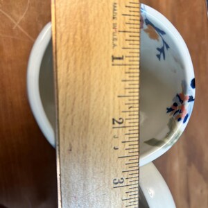 May include: A white ceramic teacup with a handle, decorated with blue, orange, and green floral patterns. The cup is approximately 3 inches (7.6 cm) tall, as measured by a wooden ruler. The text "MADE IN U.S.A." is visible on the ruler.
