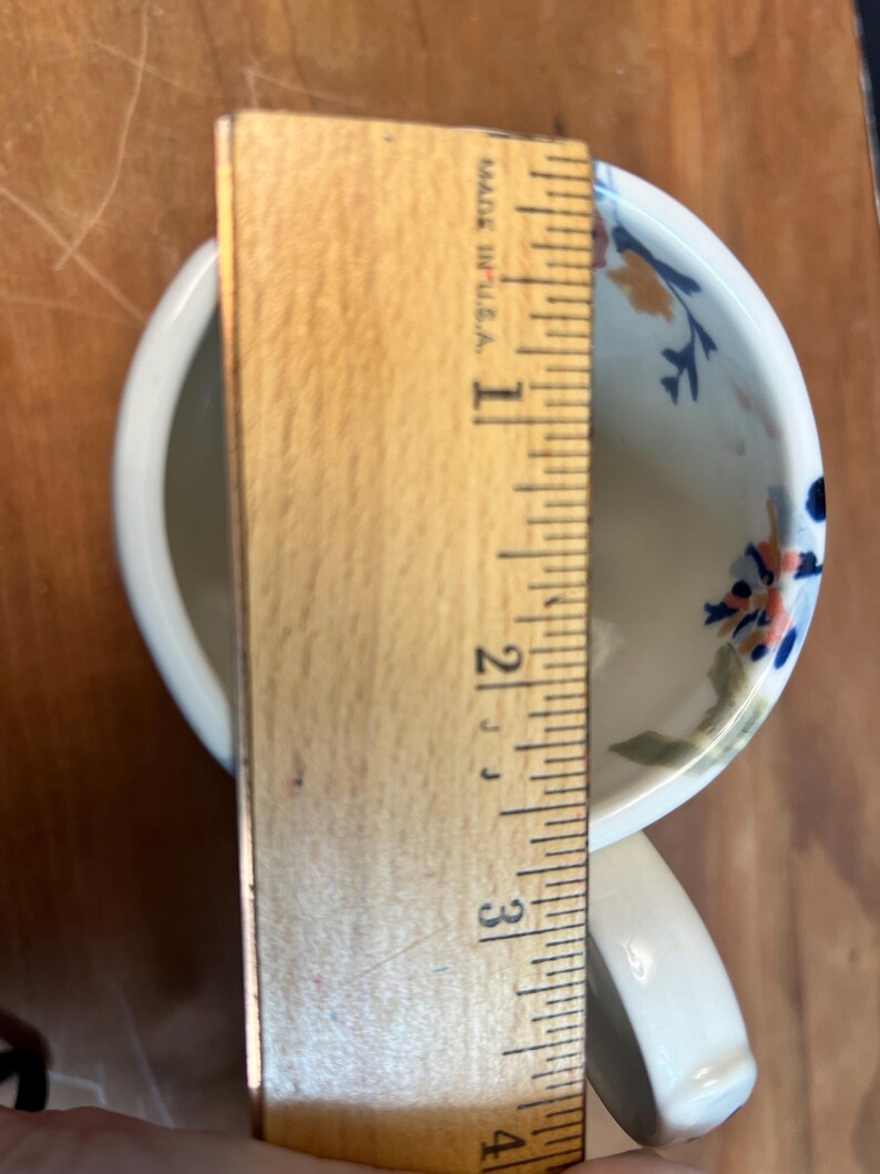 May include: A white ceramic teacup with a handle, decorated with blue, orange, and green floral patterns. The cup is approximately 3 inches (7.6 cm) tall, as measured by a wooden ruler. The text "MADE IN U.S.A." is visible on the ruler.