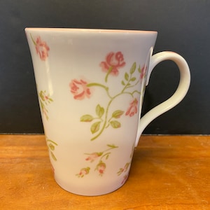 May include: A white ceramic mug with a pink and green floral pattern. The mug has a handle and a rounded shape.