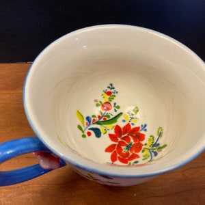 K&ouml;nnte beinhalten: Eine wei&szlig;e Keramik-Teetasse mit blauem Henkel und Rand, verziert mit einem Blumendesign in Rot, Gelb und Blau. Der Innenraum weist ein &auml;hnliches Blumenmuster auf. Ein charmantes St&uuml;ck f&uuml;r Tee oder Kaffee.