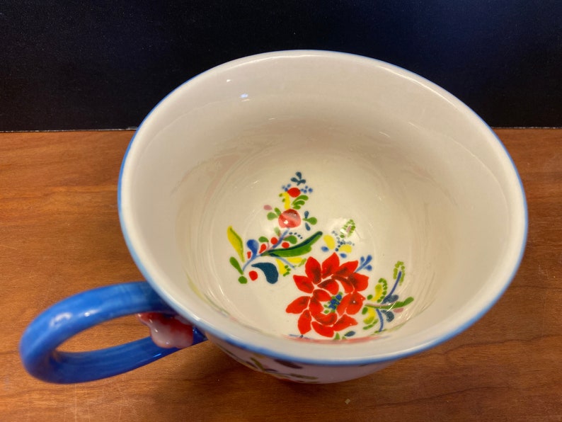 K&ouml;nnte beinhalten: Eine wei&szlig;e Keramik-Teetasse mit blauem Henkel und Rand, verziert mit einem Blumendesign in Rot, Gelb und Blau. Der Innenraum weist ein &auml;hnliches Blumenmuster auf. Ein charmantes St&uuml;ck f&uuml;r Tee oder Kaffee.