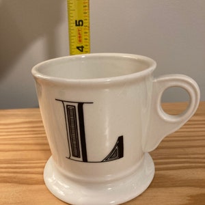 May include: White ceramic mug with a black letter L printed on the front. The mug has a handle and a wide base.