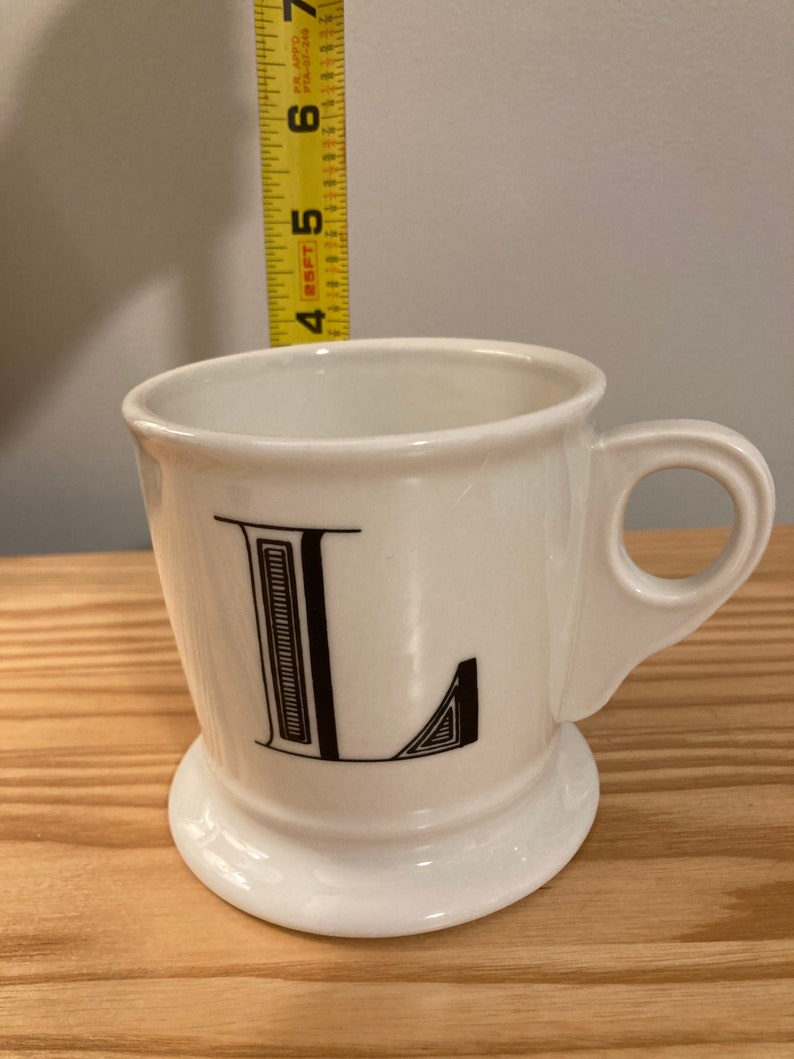 May include: White ceramic mug with a black letter L printed on the front. The mug has a handle and a wide base.
