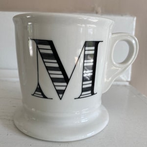 Puede incluir: Taza de cer&aacute;mica blanca con un dise&ntilde;o de la letra "M" en negro. La letra est&aacute; estilizada con l&iacute;neas horizontales. La taza tiene un asa y una base. La taza est&aacute; sobre una superficie blanca.