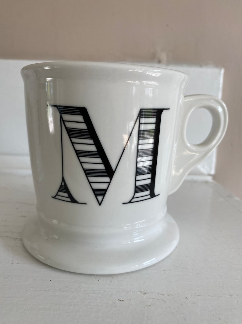 Puede incluir: Taza de cer&aacute;mica blanca con un dise&ntilde;o de la letra "M" en negro. La letra est&aacute; estilizada con l&iacute;neas horizontales. La taza tiene un asa y una base. La taza est&aacute; sobre una superficie blanca.
