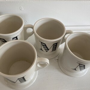 Puede incluir: Cuatro tazas de cer&aacute;mica blanca con dise&ntilde;os de letras negras. Cada taza tiene un asa y una base ancha. Las letras son en negrita, con serif. Las tazas est&aacute;n dispuestas sobre una superficie blanca.