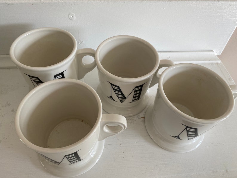 Puede incluir: Cuatro tazas de cer&aacute;mica blanca con dise&ntilde;os de letras negras. Cada taza tiene un asa y una base ancha. Las letras son en negrita, con serif. Las tazas est&aacute;n dispuestas sobre una superficie blanca.
