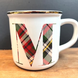 May include: White ceramic mug with a gold rim and handle. The letter "M" is prominently displayed in a plaid pattern of red, green, and black. The mug is set on a wooden surface.