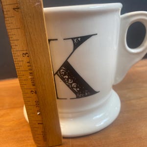 May include: White ceramic mug with a black letter "K" design. The letter is decorated with ornate details. The mug has a rounded base and a handle. A wooden ruler is placed next to the mug, showing measurements in inches.