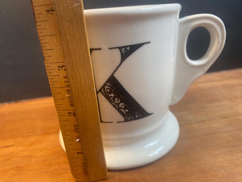 May include: White ceramic mug with a black letter "K" design. The letter is decorated with ornate details. The mug has a rounded base and a handle. A wooden ruler is placed next to the mug, showing measurements in inches.