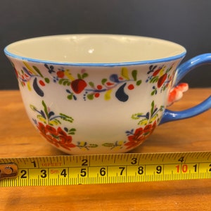 K&ouml;nnte beinhalten: Wei&szlig;e Keramik-Teetasse mit blauem Henkel und Rand, verziert mit roten, blauen und gelben Blumenmustern. Die Tasse hat einen Durchmesser von etwa 10 cm. Der Henkel hat einen kleinen, roten floralen Akzent.