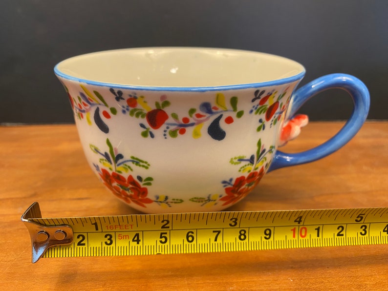 K&ouml;nnte beinhalten: Wei&szlig;e Keramik-Teetasse mit blauem Henkel und Rand, verziert mit roten, blauen und gelben Blumenmustern. Die Tasse hat einen Durchmesser von etwa 10 cm. Der Henkel hat einen kleinen, roten floralen Akzent.