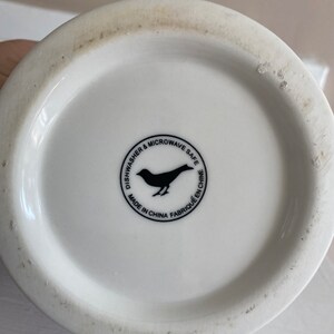 Puede incluir: Base de taza de cer&aacute;mica blanca con un logotipo de p&aacute;jaro negro y texto que dice "Dishwasher & Microwave Safe" y "Made in China Fabriqu&eacute; en Chine". La taza tiene una superficie ligeramente texturizada.