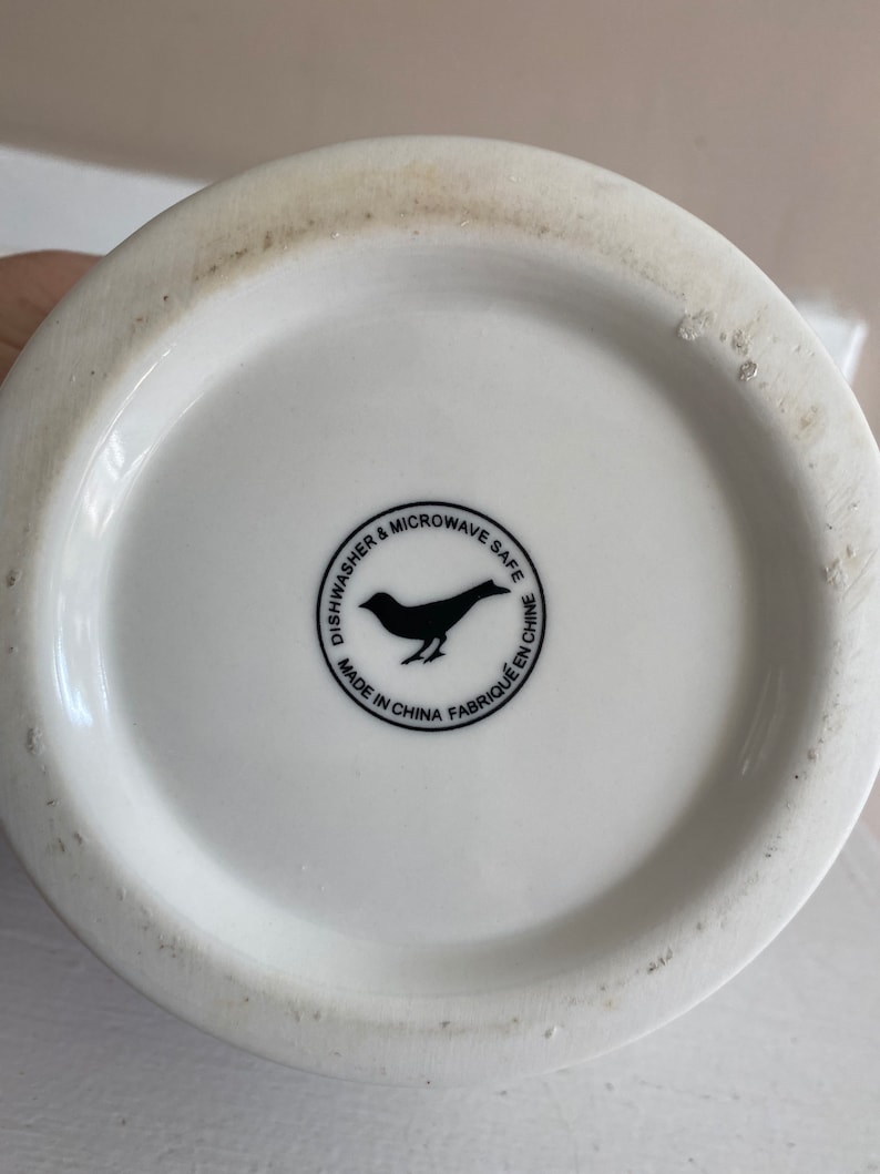 Puede incluir: Base de taza de cer&aacute;mica blanca con un logotipo de p&aacute;jaro negro y texto que dice "Dishwasher & Microwave Safe" y "Made in China Fabriqu&eacute; en Chine". La taza tiene una superficie ligeramente texturizada.