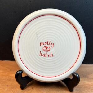 May include: A white ceramic coaster with a red border and the words "molly hatch" in red script. The coaster has a concentric circle design and is displayed on a black stand.
