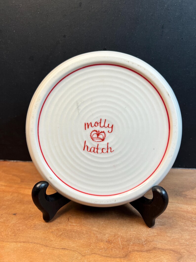 May include: A white ceramic coaster with a red border and the words "molly hatch" in red script. The coaster has a concentric circle design and is displayed on a black stand.