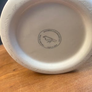 May include: Off-white ceramic vase base with a circular stamp featuring a bird illustration and the text "MICROWAVE & DISHWASHER SAFE". The stamp also includes "FABRIQU&Eacute; EN CHINE" and "MADE IN CHINA". The vase sits on a wooden surface.