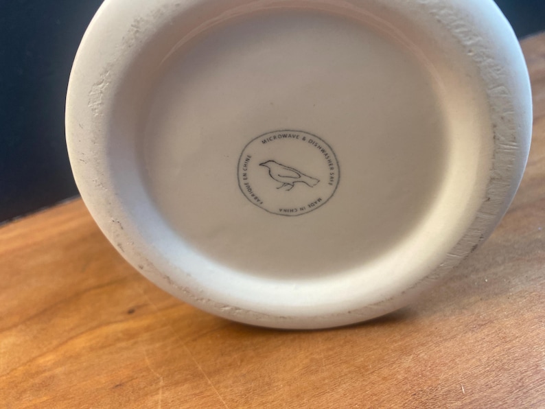 May include: Off-white ceramic vase base with a circular stamp featuring a bird illustration and the text "MICROWAVE & DISHWASHER SAFE". The stamp also includes "FABRIQU&Eacute; EN CHINE" and "MADE IN CHINA". The vase sits on a wooden surface.