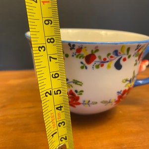 K&ouml;nnte beinhalten: Wei&szlig;e Keramik-Teetasse mit blauem Henkel und Rand, verziert mit einem Blumenmuster in Rot, Gelb und Blau. Die Tasse ist etwa 7.6 cm hoch, gemessen mit einem gelben Ma&szlig;band.