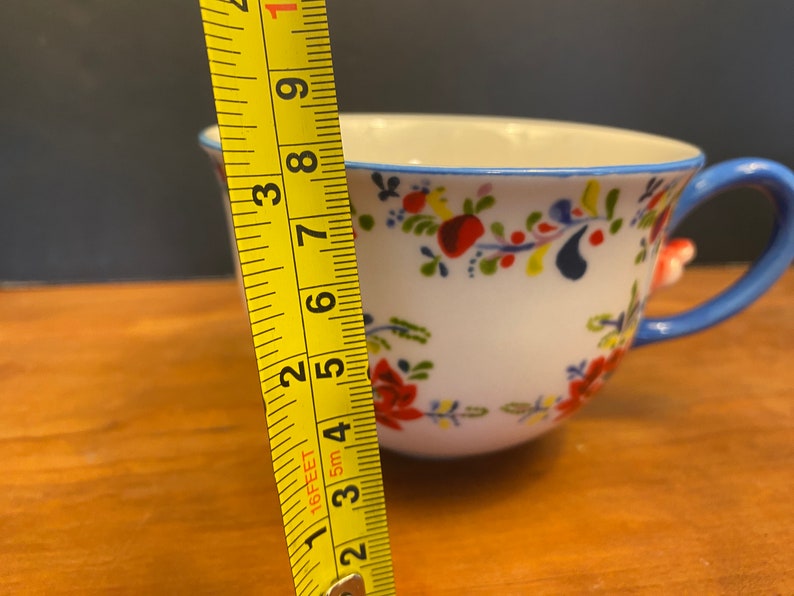 K&ouml;nnte beinhalten: Wei&szlig;e Keramik-Teetasse mit blauem Henkel und Rand, verziert mit einem Blumenmuster in Rot, Gelb und Blau. Die Tasse ist etwa 7.6 cm hoch, gemessen mit einem gelben Ma&szlig;band.