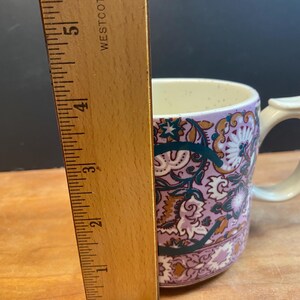 May include: A ceramic mug with a floral pattern in shades of purple, teal, and gold. The mug has a cream-coloured handle and interior. The ruler next to the mug shows a height of approximately 10 cm.