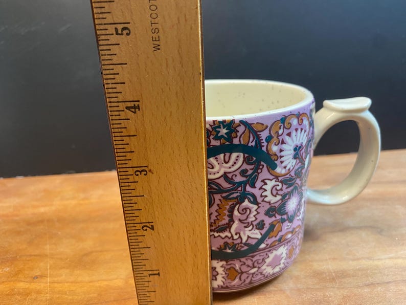 May include: A ceramic mug with a floral pattern in shades of purple, teal, and gold. The mug has a cream-coloured handle and interior. The ruler next to the mug shows a height of approximately 10 cm.