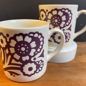 May include: Two white ceramic mugs with purple floral designs. The mugs have a handle and are in focus.