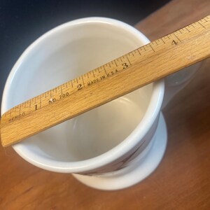 May include: A white ceramic shaving mug with a pedestal base, featuring a brown floral design. A wooden ruler, marked "Made in U.S.A.", rests across the mug's opening, measuring approximately 5 inches.