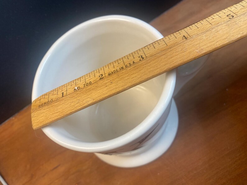 May include: A white ceramic shaving mug with a pedestal base, featuring a brown floral design. A wooden ruler, marked "Made in U.S.A.", rests across the mug's opening, measuring approximately 5 inches.