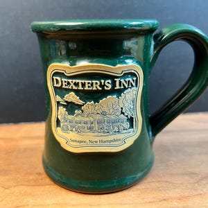 May include: A dark green ceramic mug featuring a raised, tan-colored emblem on the front. The emblem displays the text "DEXTER'S INN" above a relief illustration of a building and trees, with the text "Sunapee, New Hampshire" at the bottom.