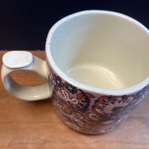 May include: A ceramic mug with a distinctive handle. The mug showcases a floral pattern in purple, brown, and teal hues. The interior is a light cream colour, and the handle has a small, rectangular accent. Suitable for hot or cold drinks.