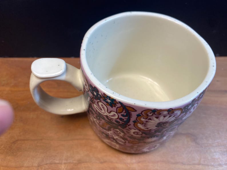 May include: A ceramic mug with a distinctive handle. The mug showcases a floral pattern in purple, brown, and teal hues. The interior is a light cream colour, and the handle has a small, rectangular accent. Suitable for hot or cold drinks.