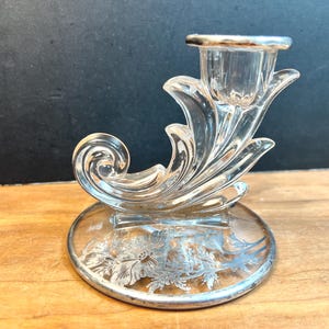 May include: Clear glass candlestick holder with a silver rim and base. The base features a floral design. The candlestick has a decorative, swirling design and is set on a wooden surface.