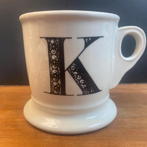 May include: White ceramic mug with a black letter "K" on the front. The letter is decorated with a detailed, ornate design. The mug has a handle and a wide, stable base. The mug is on a wooden surface.