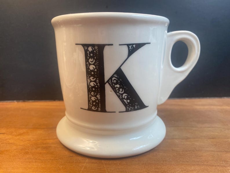 May include: White ceramic mug with a black letter "K" on the front. The letter is decorated with a detailed, ornate design. The mug has a handle and a wide, stable base. The mug is on a wooden surface.