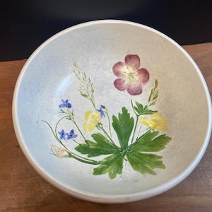 Może przedstawiać: Okrągła, kremowa ceramiczna miska z kwiatowym wzorem. Ręcznie malowane kwiaty obejmują duży, ciemnoróżowy kwiat, żółte i niebieskie kwiaty oraz zielone liście. Misa ma lekko teksturowaną powierzchnię.