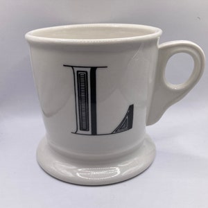 May include: White ceramic mug with a black letter L on the front. The mug has a wide base and a handle.