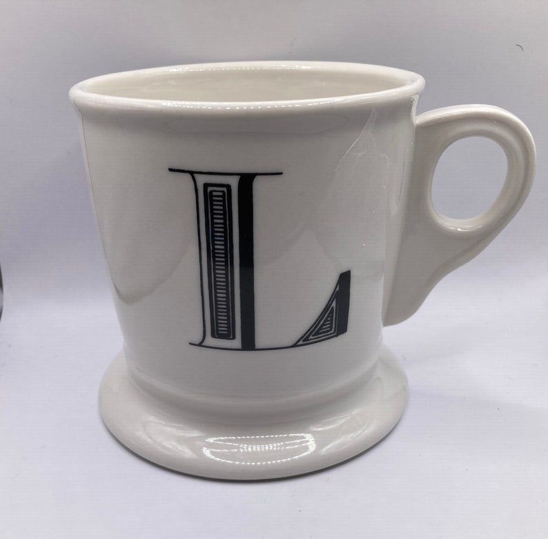 May include: White ceramic mug with a black letter L on the front. The mug has a wide base and a handle.