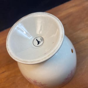 K&ouml;nnte beinhalten: Ein wei&szlig;er Keramik-Eierbecher mit ausgestelltem Fu&szlig; und rundem K&ouml;rper. Der Becher hat ein kleines, kreisf&ouml;rmiges schwarz-wei&szlig;es Logo auf der Basis. Der K&ouml;rper des Bechers hat ein zartes Blumenmuster in Rosa.