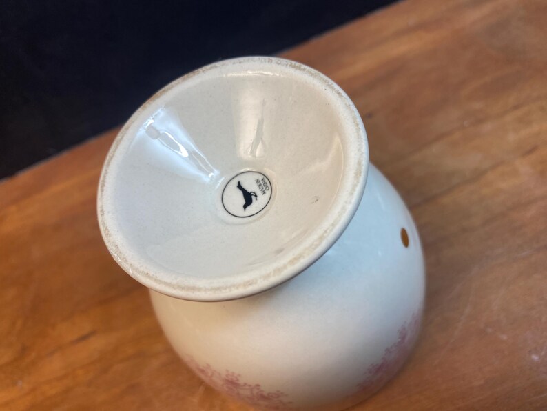 K&ouml;nnte beinhalten: Ein wei&szlig;er Keramik-Eierbecher mit ausgestelltem Fu&szlig; und rundem K&ouml;rper. Der Becher hat ein kleines, kreisf&ouml;rmiges schwarz-wei&szlig;es Logo auf der Basis. Der K&ouml;rper des Bechers hat ein zartes Blumenmuster in Rosa.